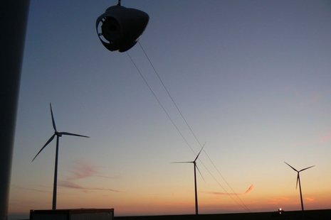Eine Rotornabe wird bei Sonnenuntergang in die Luft gehoben. Im Hintergrund sind drei Windkraftanlagen zu sehen.