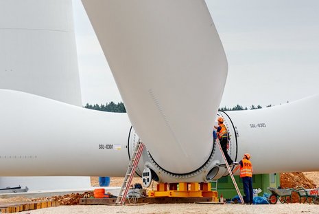 Montage der Rotorblätter an der Windkraftanlage. Zwei Arbeiter koordinieren den Vorgang.