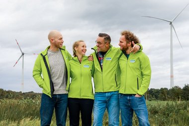 Vier Menschen stehen vor einer Windenergie-Anlage und schauen sich lachend an. Sie tragen hellgrüne Jacken mit dem Logo von Green Planet Energy.